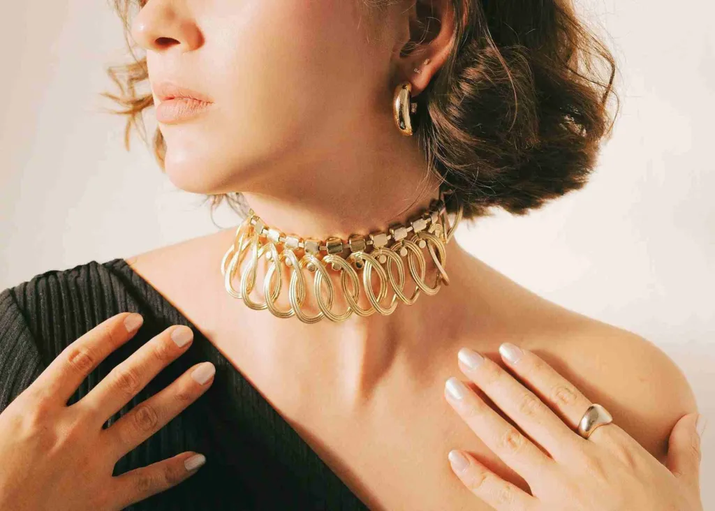 A close-up of a person wearing an ornate gold choker and hoop earrings, with hands delicately resting on their shoulders.