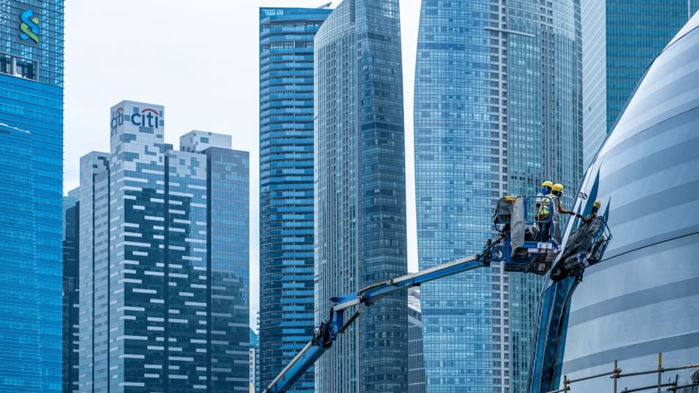 Two workers on a lift inspecting a curved facade of a building in Singapore with skyscrapers in the background, indicating the need of skilled Singapore Construction Labor Market.