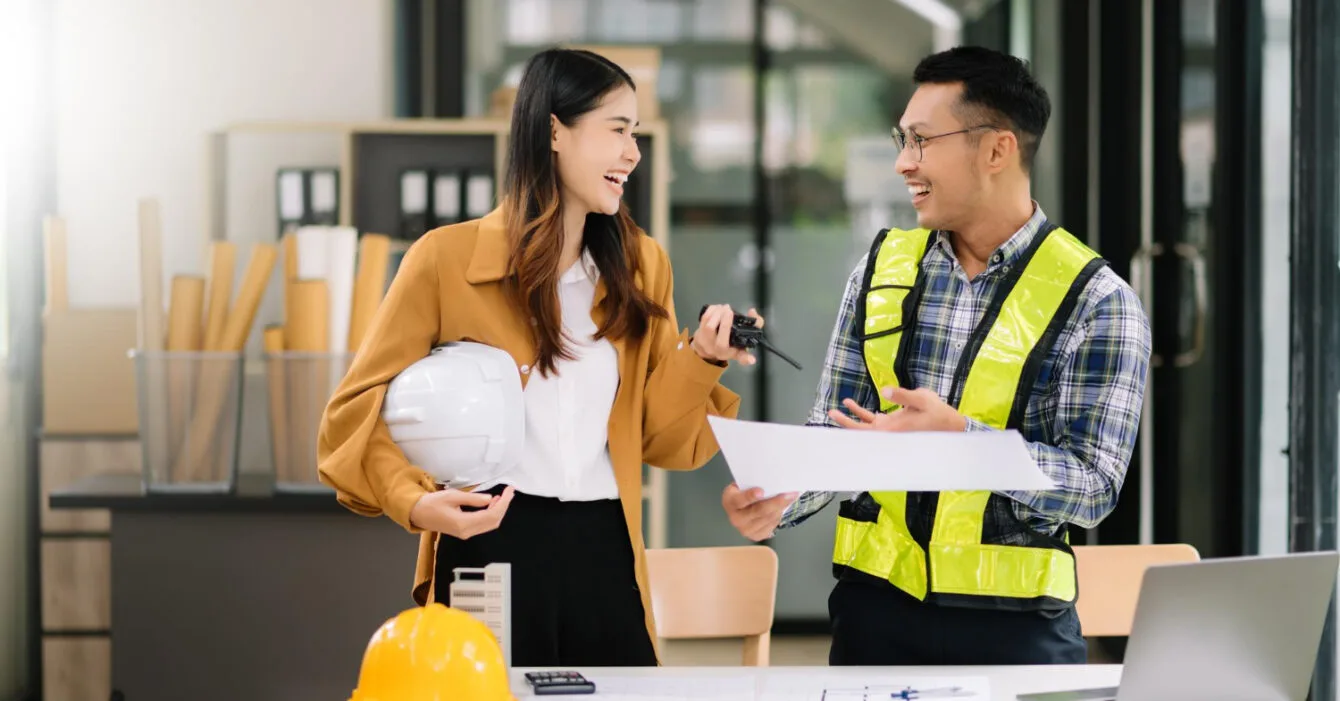 Singapore Public-Private Partnership Models - A woman holds a hard hat while reviewing documents with a man in a safety vest at a modern office table filled with construction plans.