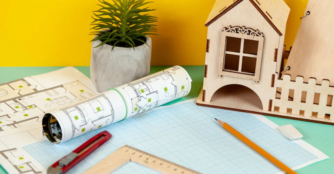 Singapore Sustainable Materials Adoption: Model house with architectural blueprints, a ruler, pencil, and plant on a two-tone background.