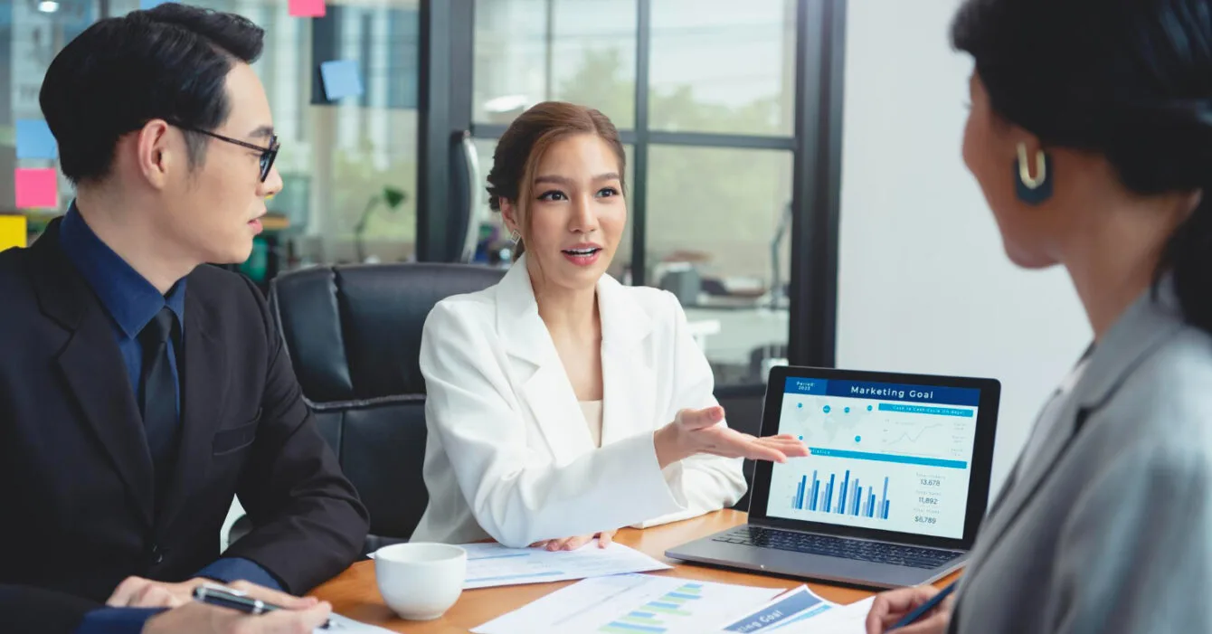 A business meeting in a modern office, with a woman presenting marketing data on a laptop to two colleagues to show Singapore Business Environment Trends.